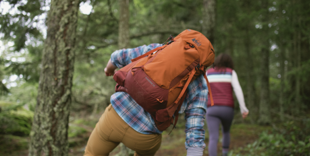 Two people are walking through a forest. One person has an orange backpack on.